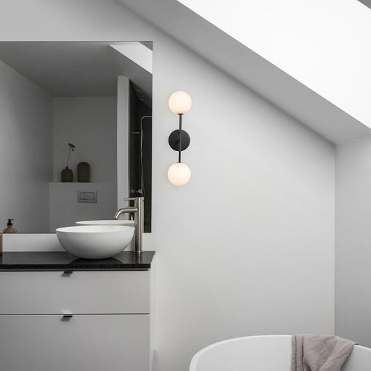 Modern badrumsbelysning med vägglampor i svart och varmvit ton, elegant spegel och stilren inredning.