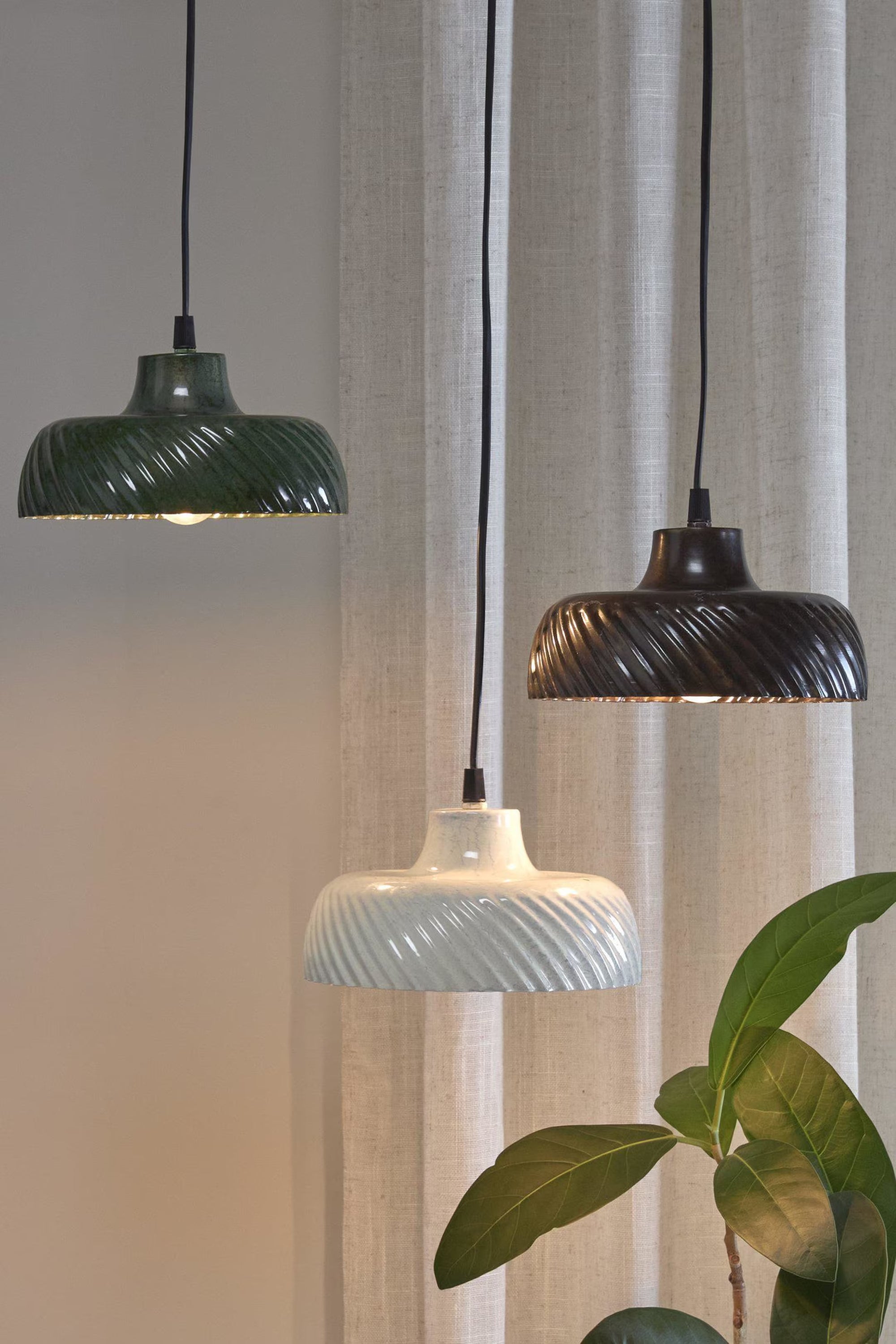 Tre keramiska fönsterlampor i grönt, brunt och vitt hängande framför ett fönster med ljusa gardiner och en grön växt i förgrunden.
