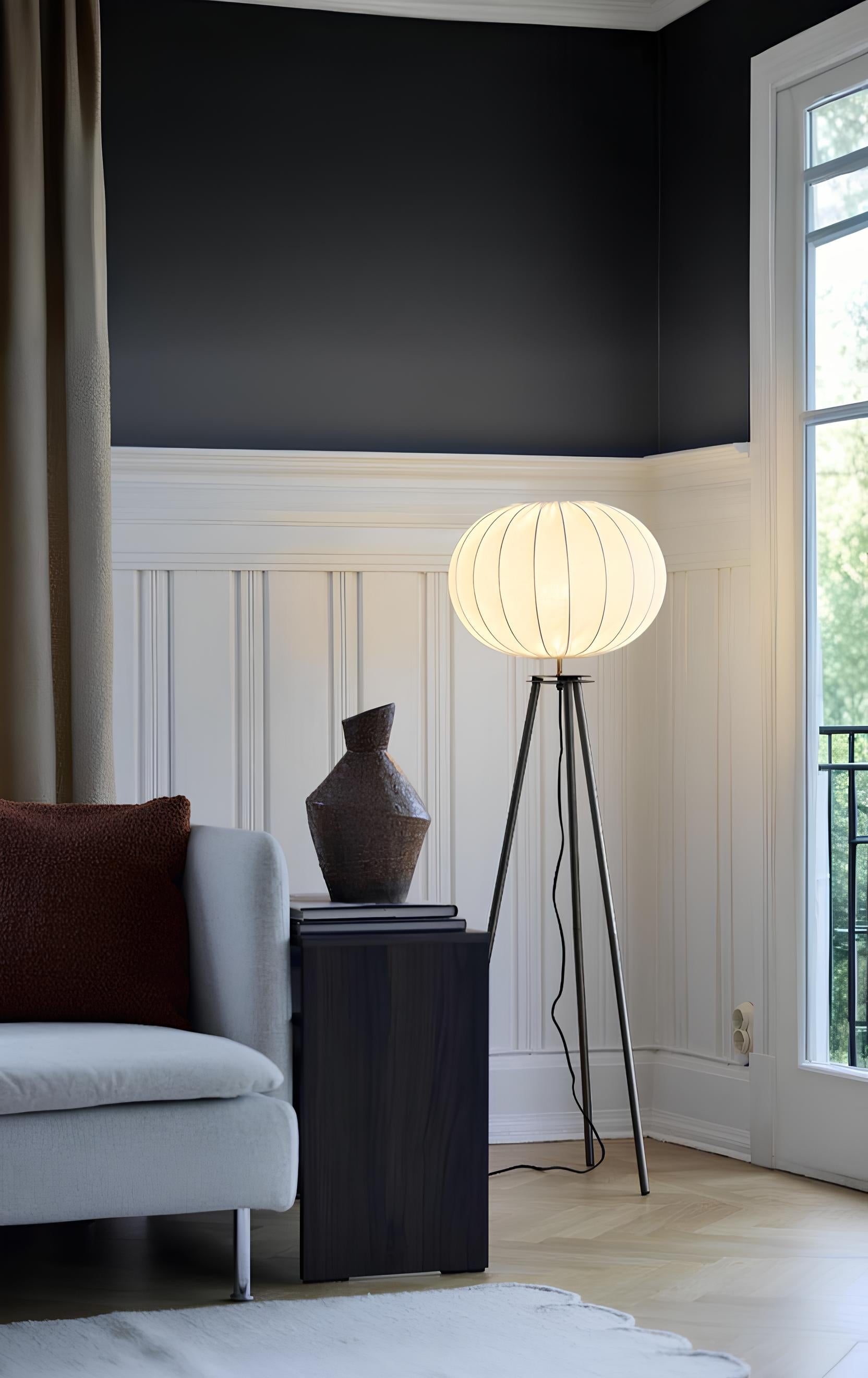 Golvlampa med rund, vit lampskärm på trebent stativ placerad i ett elegant hörn av vardagsrummet med soffa, sidobord och naturligt ljusinsläpp.