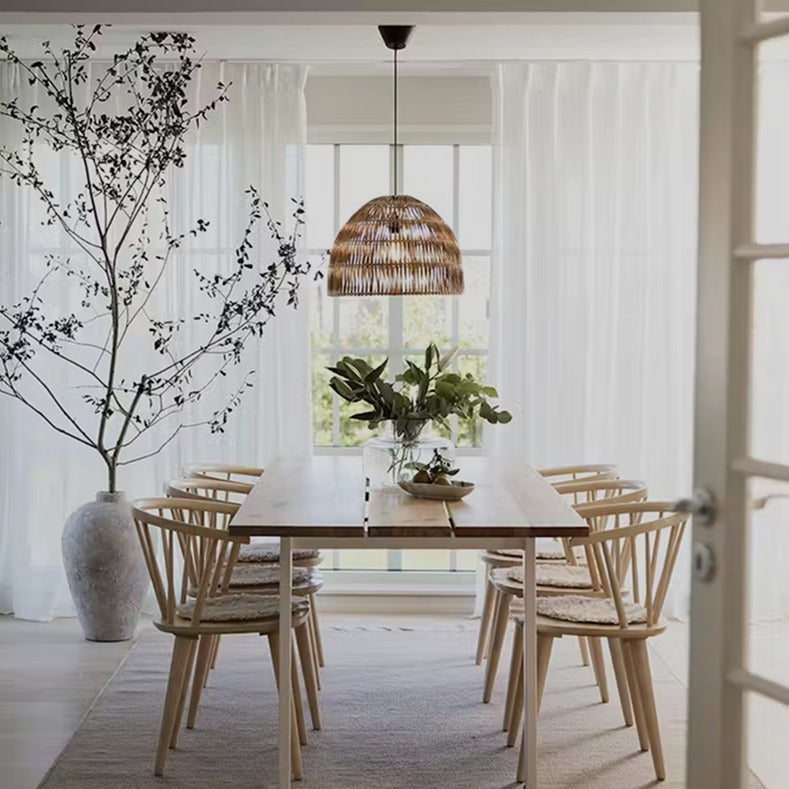 Ljus matsal med naturinspirerad taklampa i rotting ovanför ett träbord med sex stolar och gröna växter som dekoration.