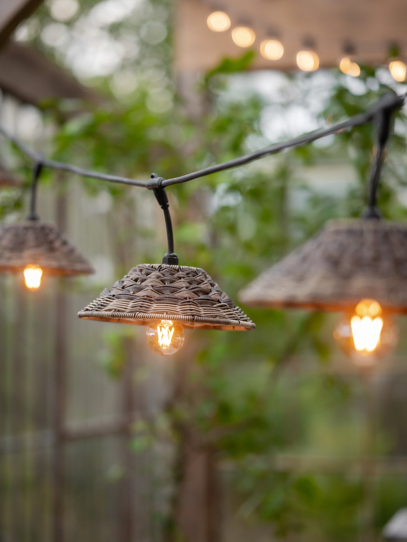 Ljusslinga med flätade lampskärmar i rottingstil hängande utomhus, med varmvitt sken och grön växtlighet i bakgrunden.