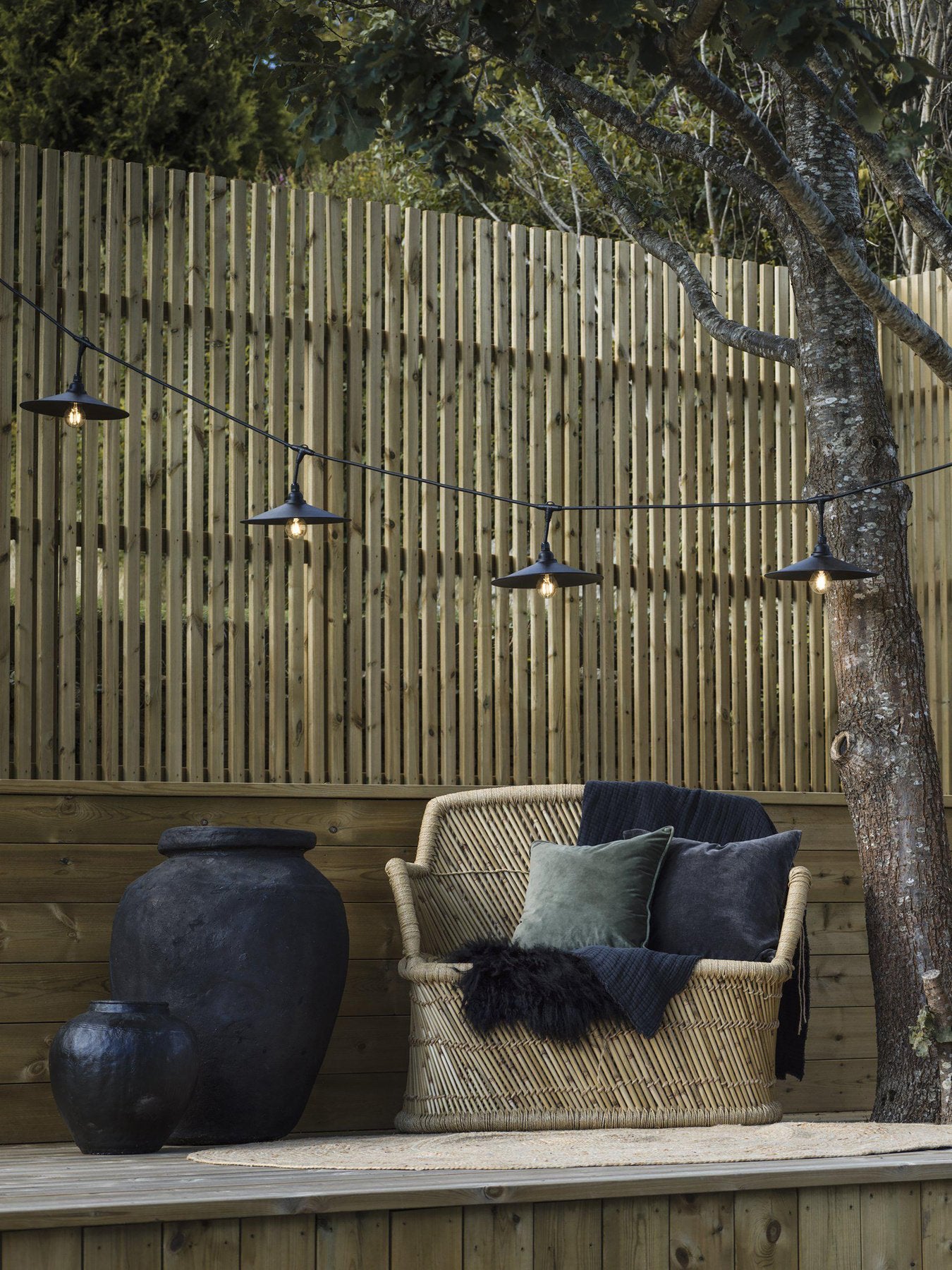 Svart ljusslinga med breda skärmar upphängd mellan träd och staket ovanför en utomhussittplats med rottingsoffa, kuddar och stora dekorationskrukor i träinramad miljö.