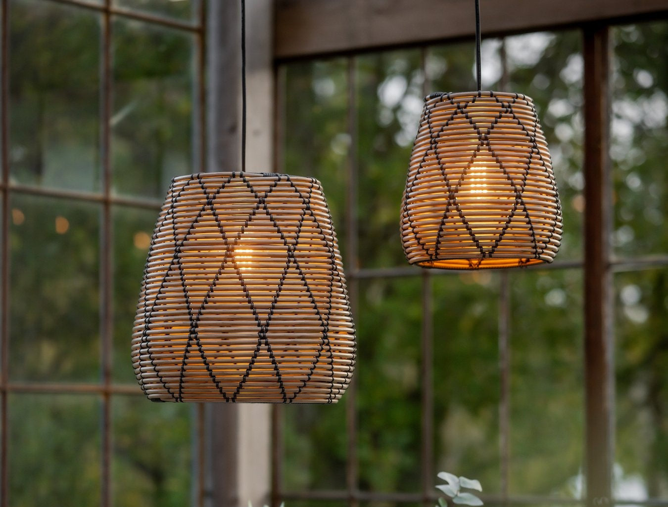Två rottinglampor hängande framför stora fönster med utsikt över grönska. Skapar ett varmt och stämningsfullt ljus.