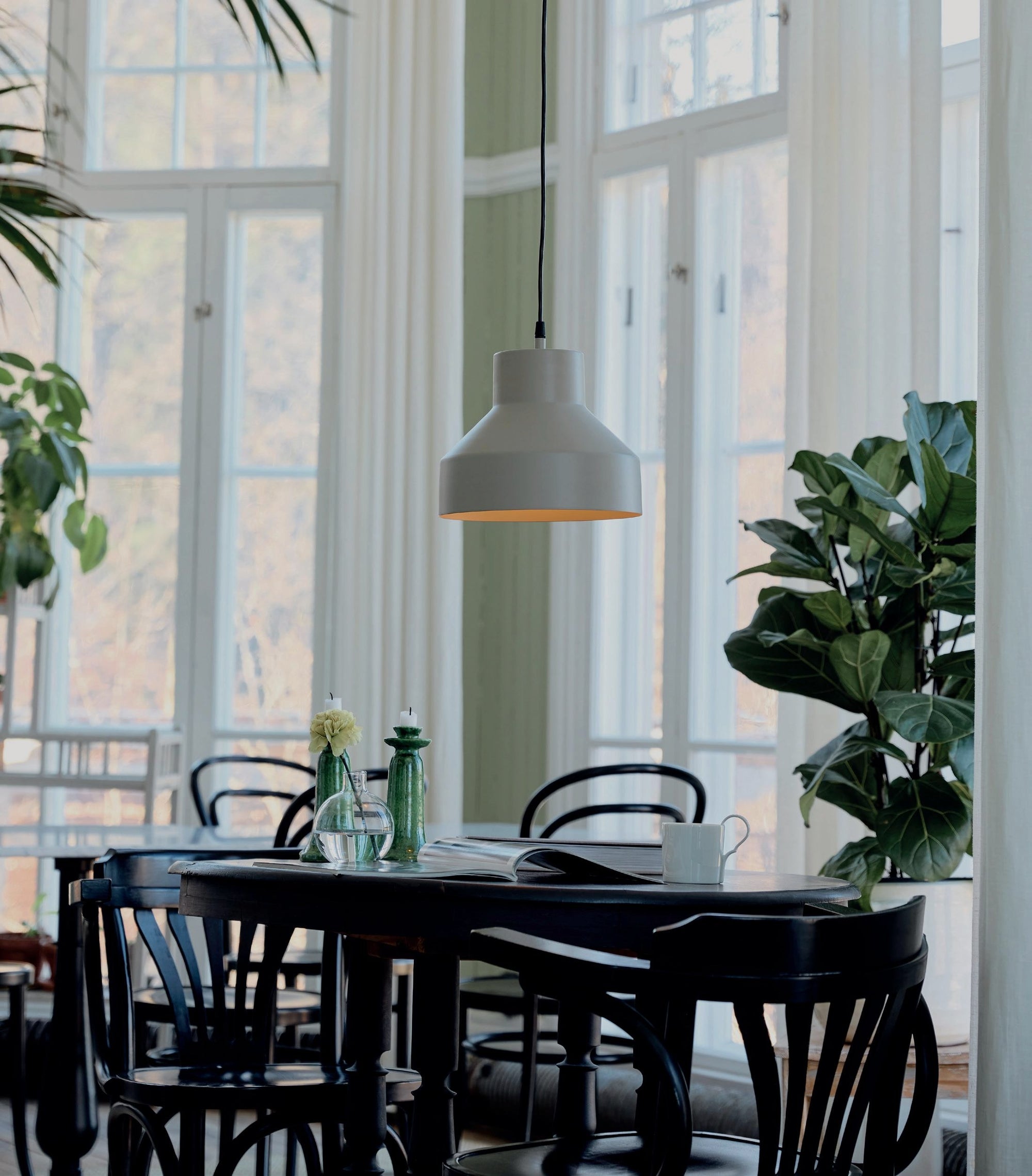 Modern taklampa ovanför ett svart matbord i ett ljust rum med stora fönster och gröna växter.