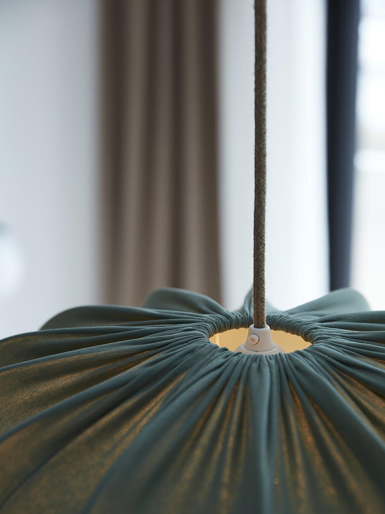 Närbild på upphängningen av en taklampa med veckad mörkgrön textilskärm och textilklädd sladd, mot suddig bakgrund med gardin och fönster.
