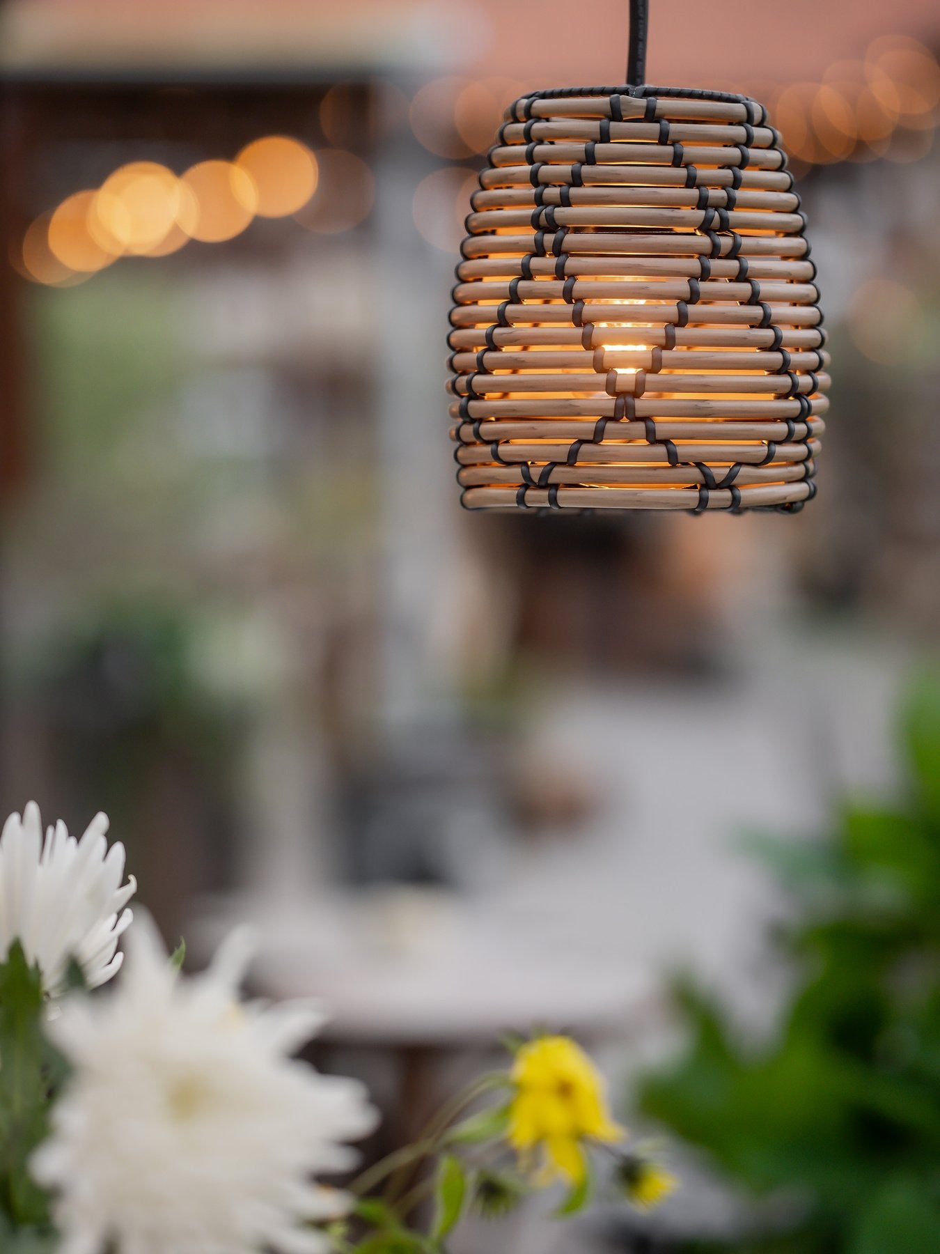 Närbild på en tänd taklampa i rottingdesign hängande utomhus, med suddiga ljusslingor och blommor i förgrunden.