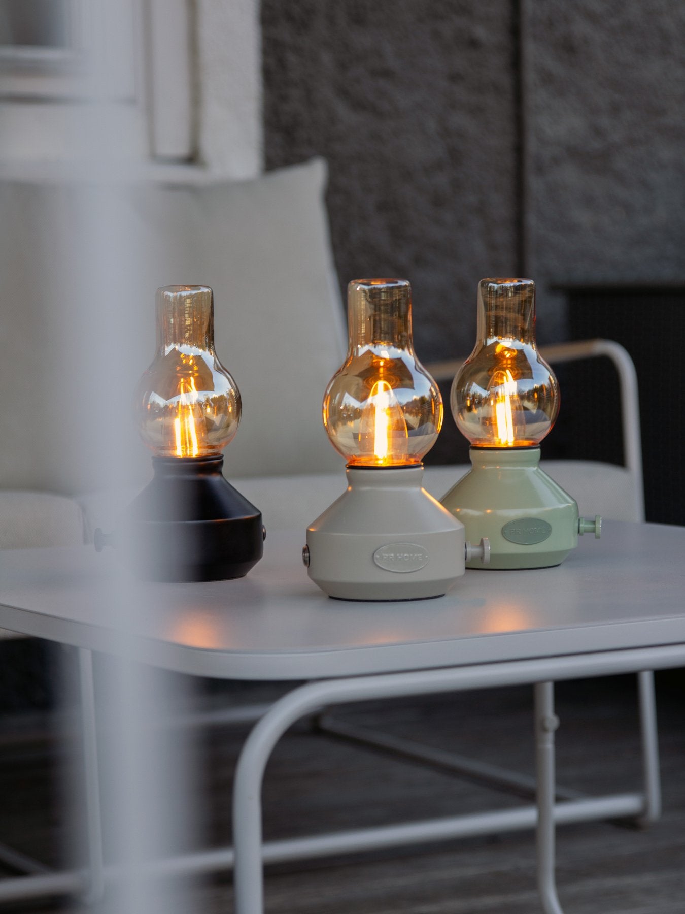 Tre uppladdningsbara bordslampor i svart, beige och ljusgrön färg står tända på ett vitt utomhusbord mot en mörk fasad.