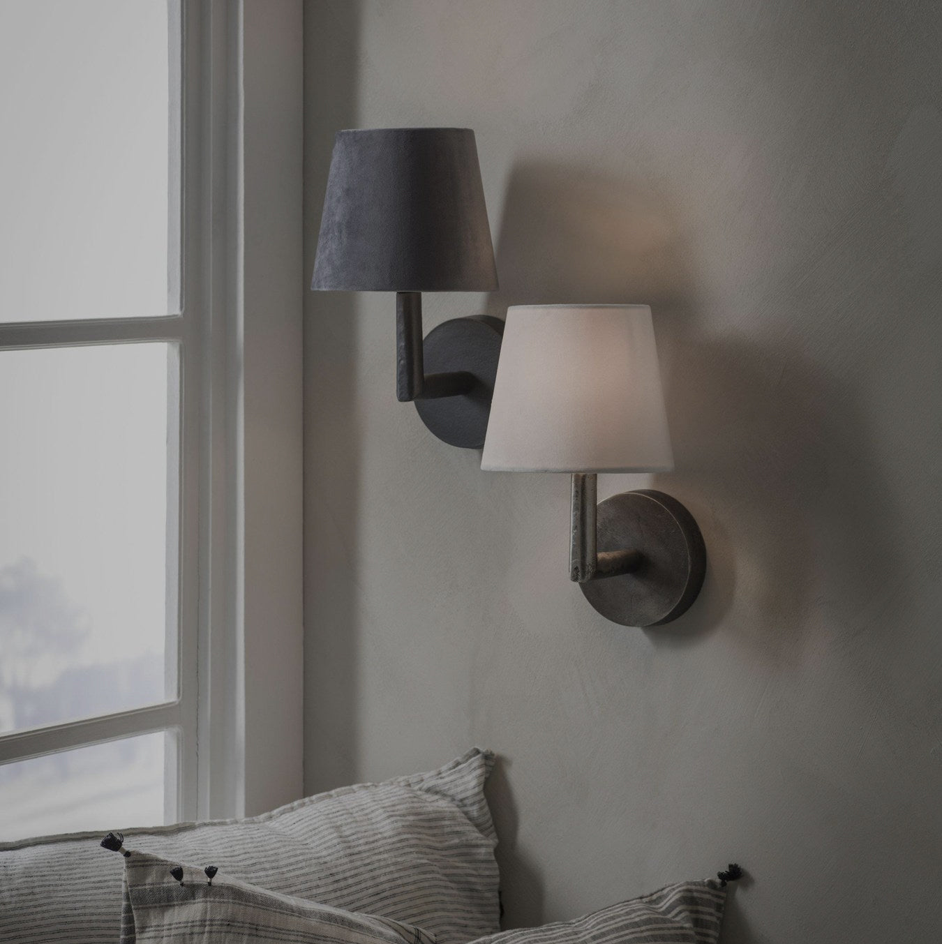 Svart vägglampa monterad på grå vägg ovanför soffa med kudde, intill ett fönster med naturligt ljusinsläpp.